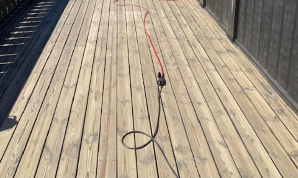 Bilde av en nylig rengjort terrasse med lysere treplanker. En rød strømkabel ligger på terrassen, som ser ren og klar ut for videre behandling eller bruk.