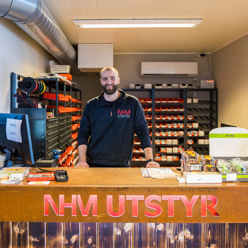 En smilende butikkmedarbeider står bak disken i en NHM Utstyr-butikk, omgitt av organisert verktøy og utstyr, klar til å hjelpe kunder.