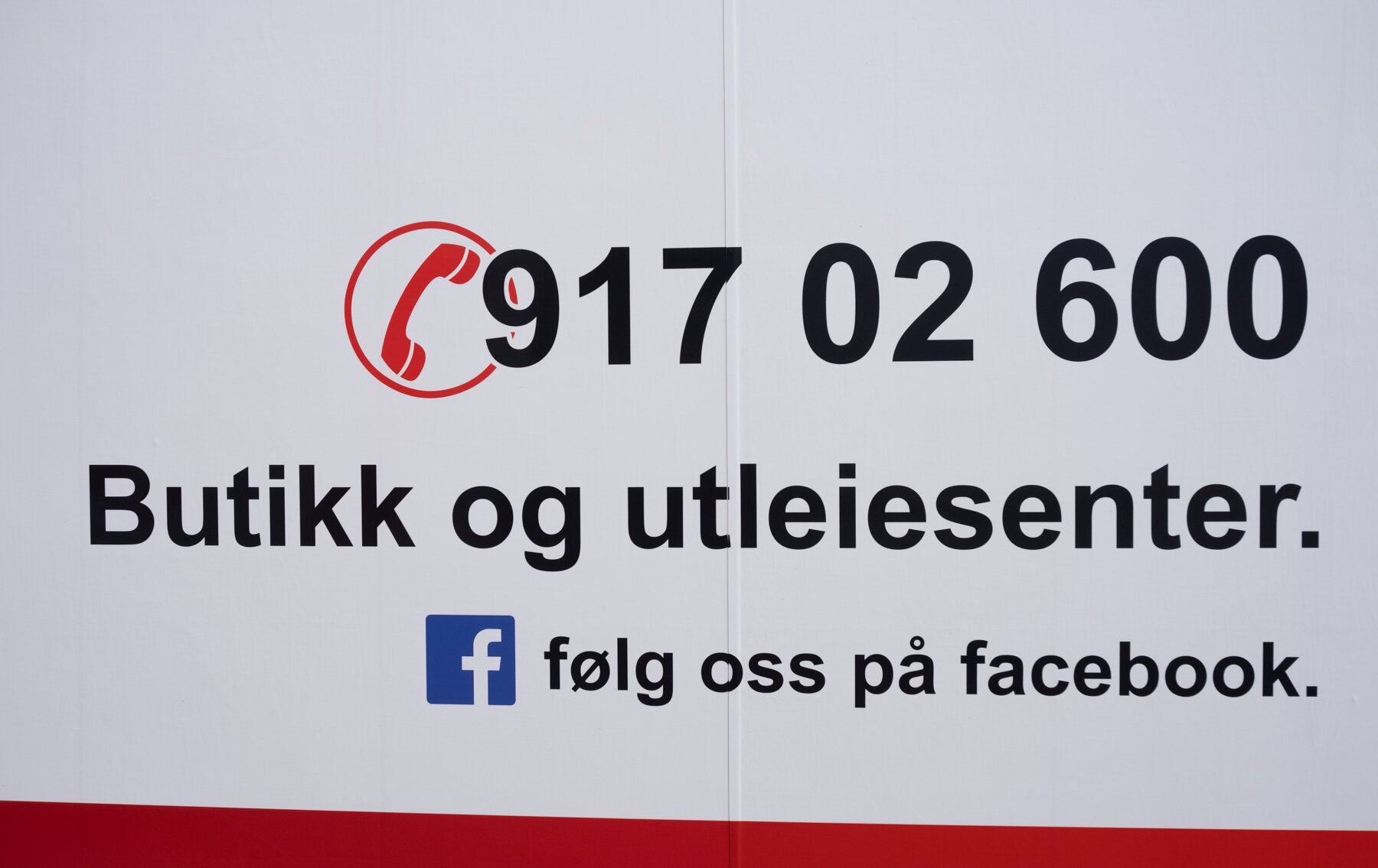 Skilt for butikk og utleiested med telefonnummer 917 02 600 og oppfordring til å følge dem på Facebook. Skiltet har et rødt telefonikon og svart tekst på hvit bakgrunn.