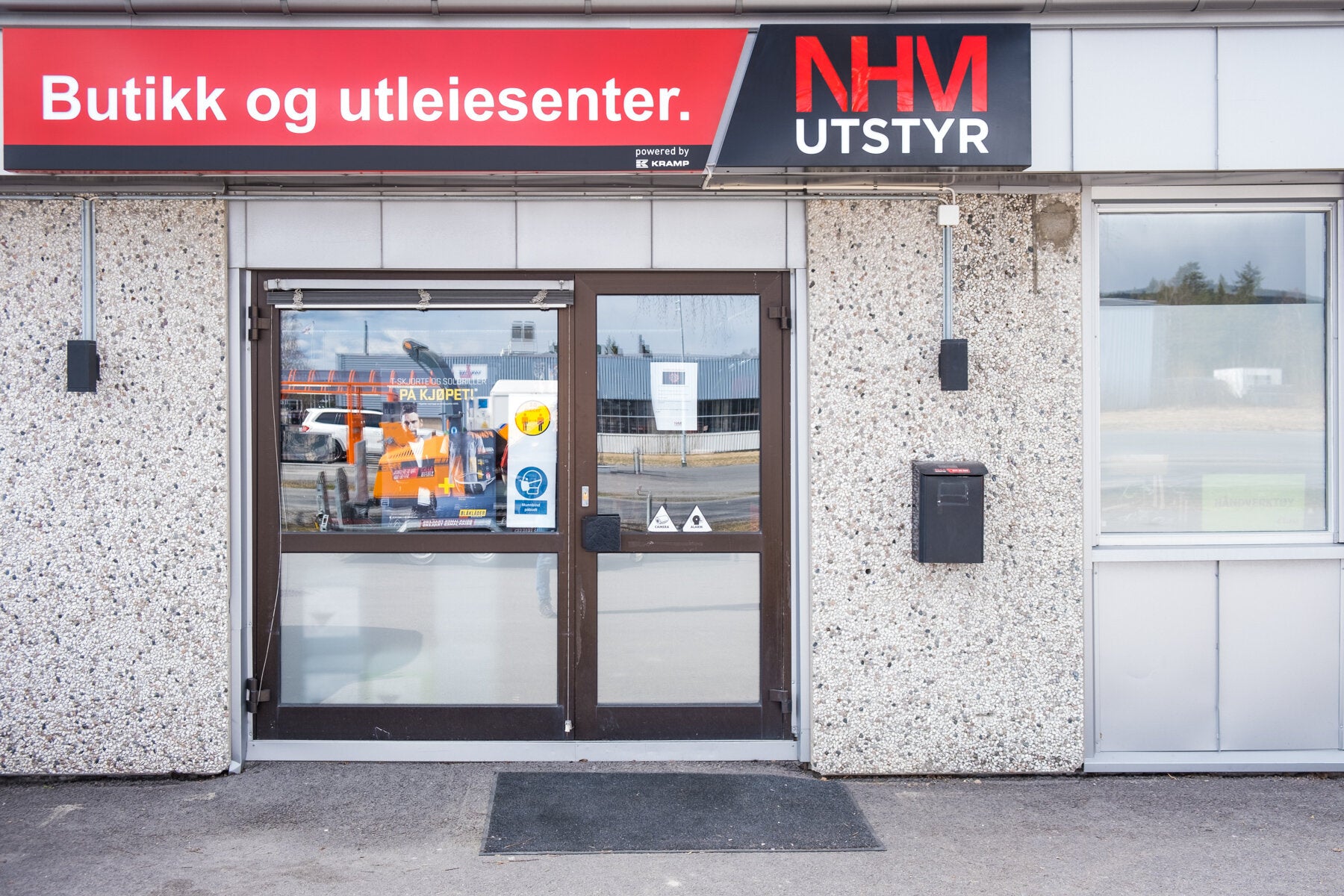 Inngang til NHM Utstyr butikk og utleiesenter med skilt over døren. Reklameplakater på døren og vinduet, samt en postkasse ved siden av inngangen.