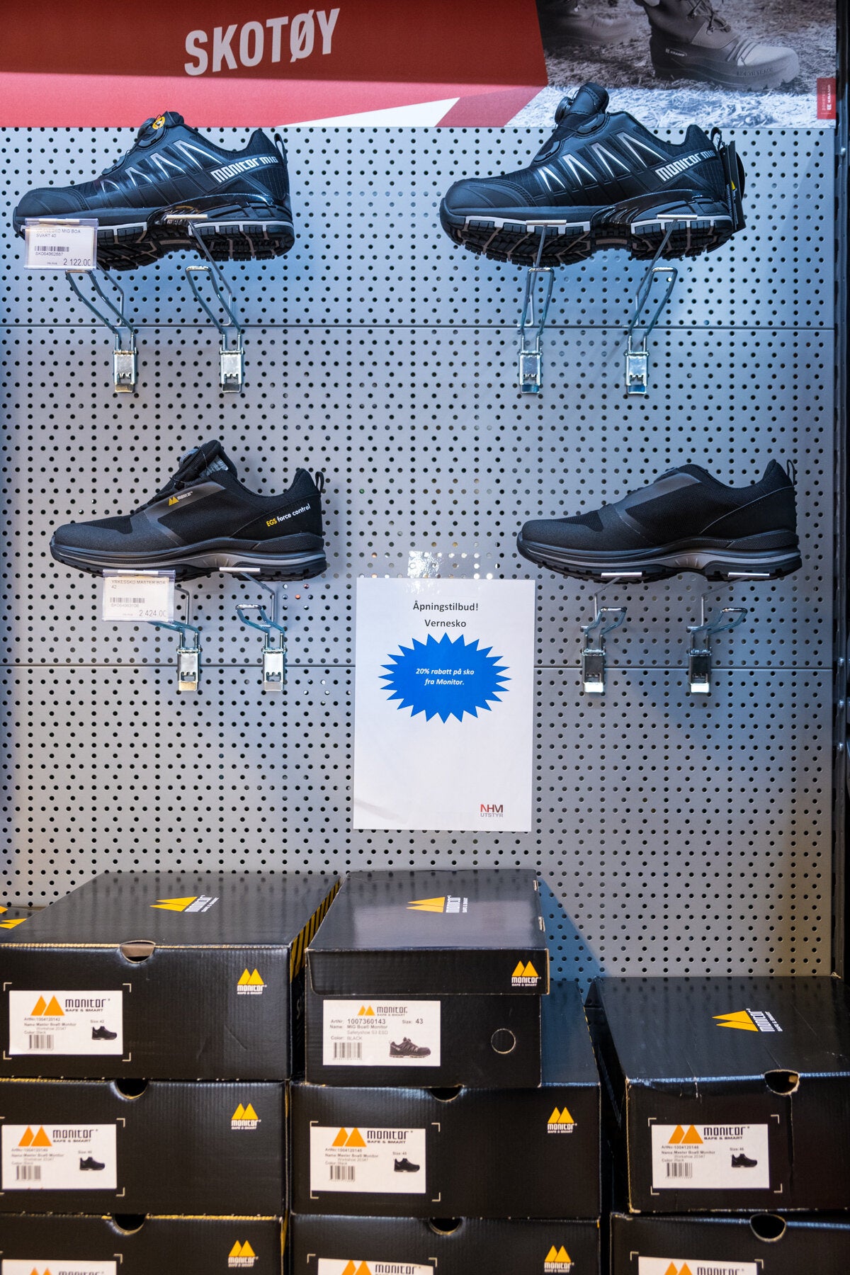 En butikkseksjon merket "SKOTØY" viser frem vernesko på hyller og i esker, med et skilt som reklamerer for et åpningssalgetilbud med 20% rabatt på utvalgte sko.