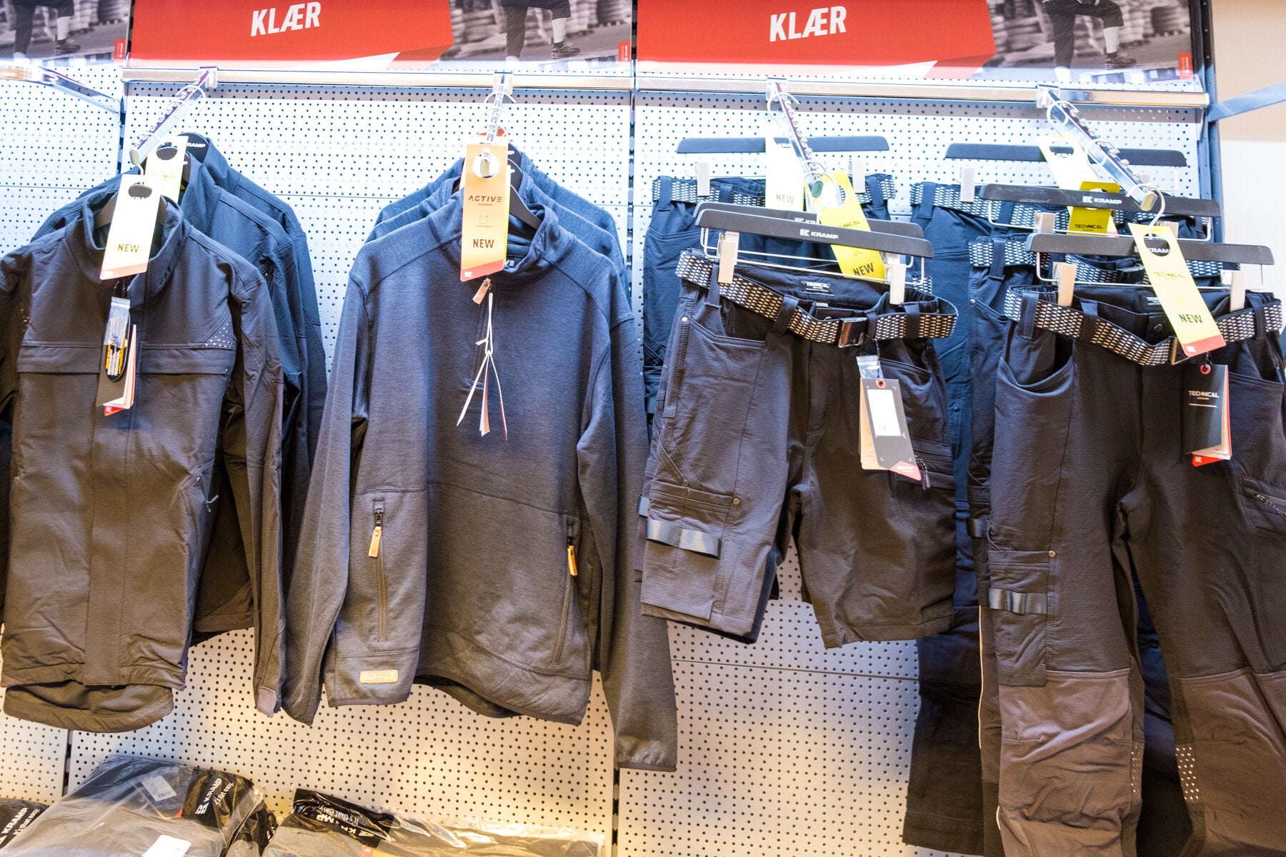 Arbeidsklær hengt opp pent i en butikk, inkludert jakker og arbeidsbukser med belter, klare for kjøp. Over seksjonen er det et skilt som sier "KLÆR."
