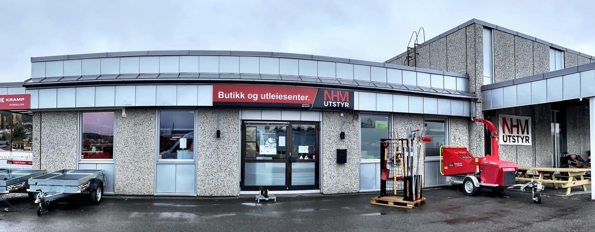 Bilde av NHM Utstyr butikk og utleiested. Viser fronten av bygningen med utleieutstyr som tilhengere og hageredskap. Skilt og logoer er godt synlige.